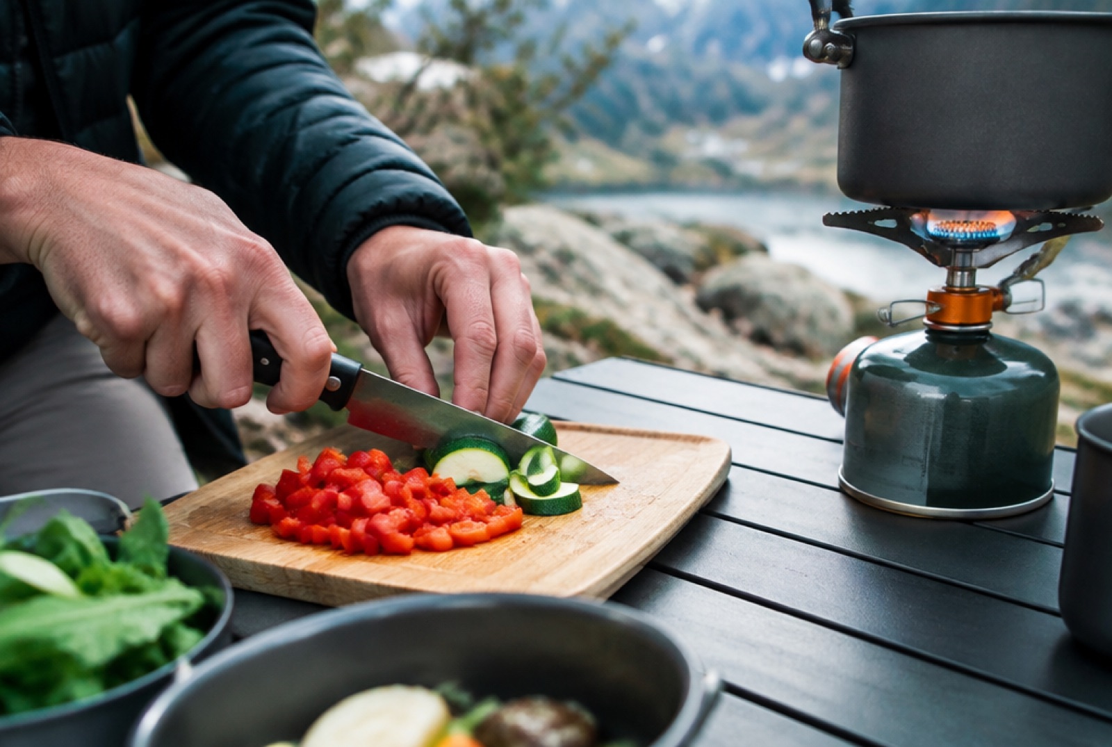 Nahaufnahme von Händen, die Gemüse neben einem brennenden Campingkocher schneiden