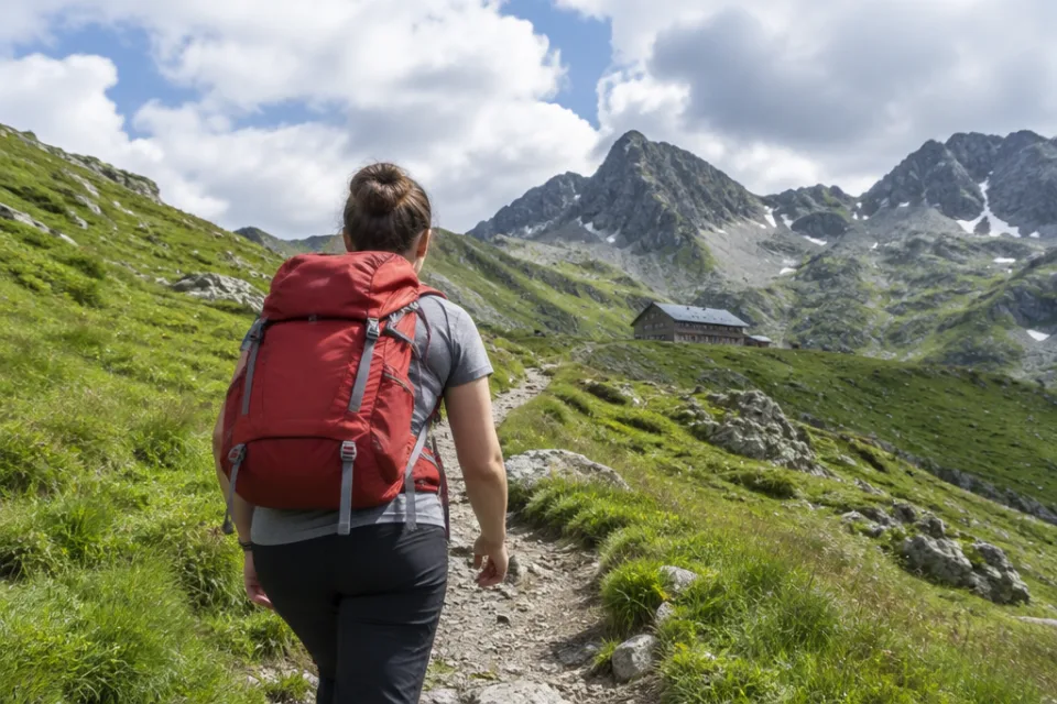 Wanderer mit rotem Tagesrucksack auf einem Bergpfad Richtung Berghütte